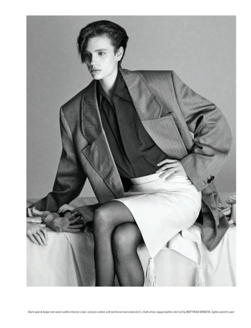 Fashion editorial shot: an androgynous model in an oversized grey blazer and white skirt sits with their hand resting on a person's hand.