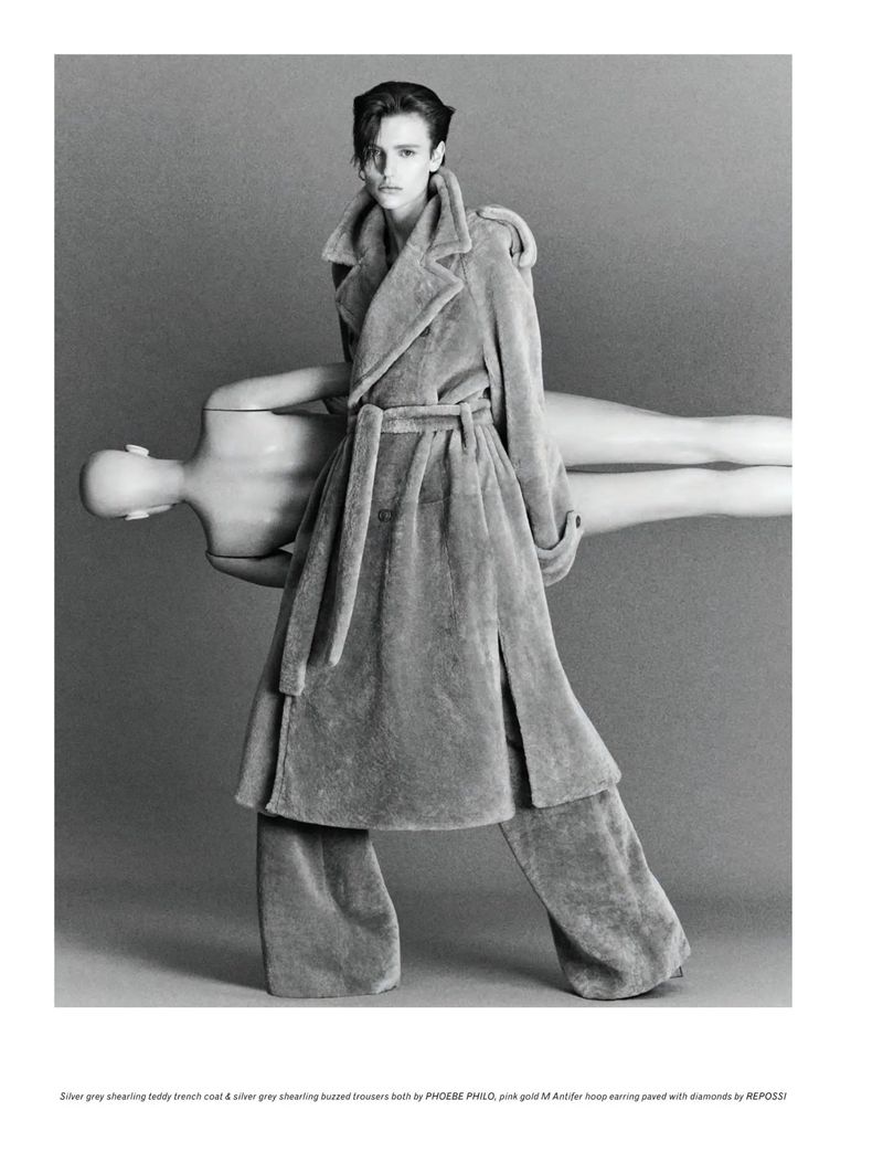 Woman in shearling coat and pants holding a mannequin in a studio setting with gray background in a fashion editorial style photograph.