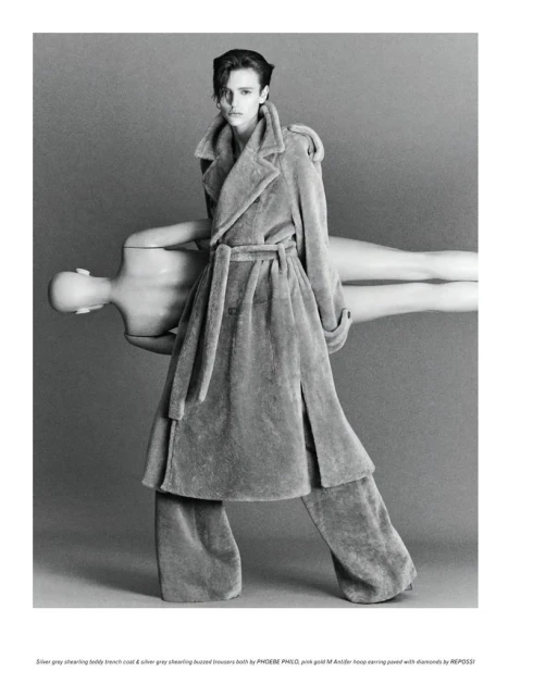 Woman in shearling coat and pants holding a mannequin in a studio setting with gray background in a fashion editorial style photograph.