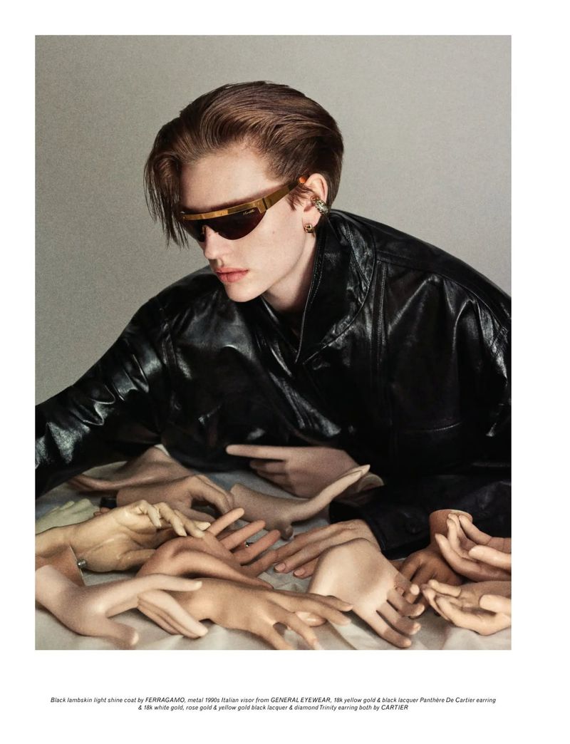 Person with slicked-back auburn hair poses wearing sunglasses and a black leather jacket surrounded by disembodied hands.