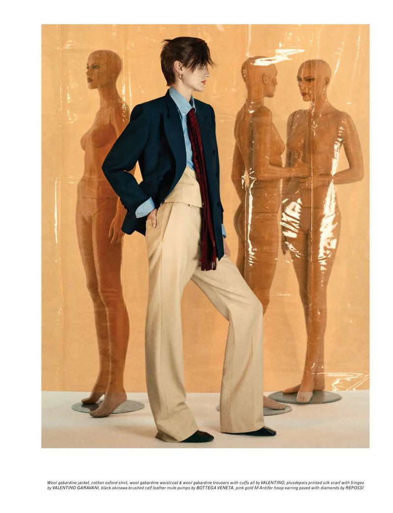 Woman in tailored blazer, button-down shirt, trousers, and scarf posing in front of mannequins wrapped in clear plastic film.