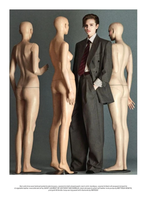 A fashionable adult in a grey suit stands between three nude mannequins against a grey backdrop in a studio lighting setup.