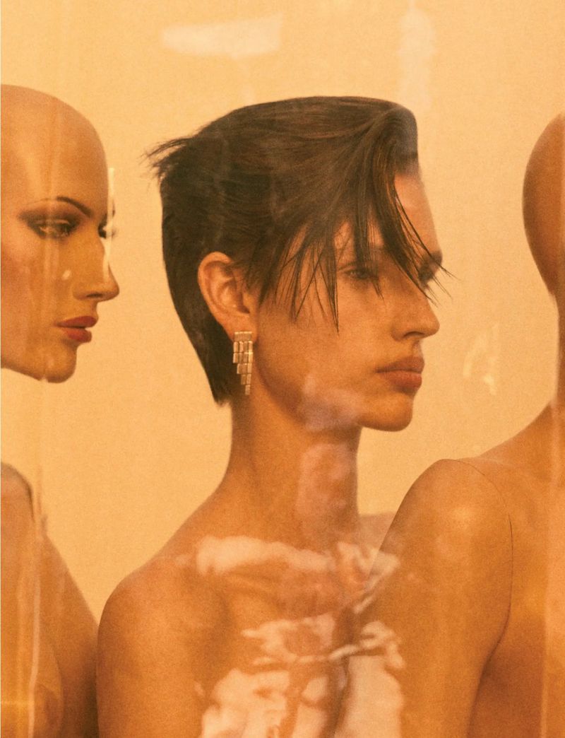 Portrait of a person with short dark hair standing near two mannequin heads, all positioned in a bright orange environment with a glossy finish.