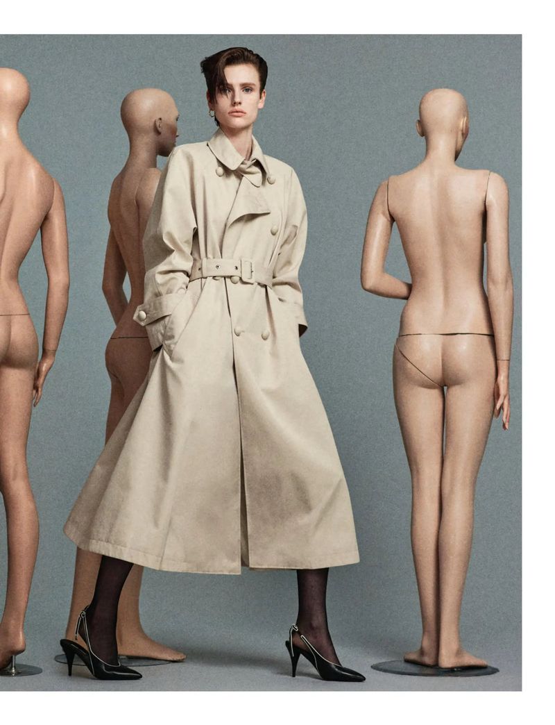 Fashion shot of a woman in a trench coat surrounded by nude mannequins in a studio setting with grey background. Editorial style.