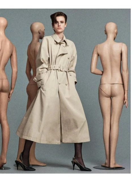Fashion shot of a woman in a trench coat surrounded by nude mannequins in a studio setting with grey background. Editorial style.