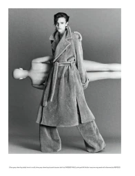 Model wearing a textured belted coat and wide trousers standing before a plain seamless background in a monochrome studio portrait.