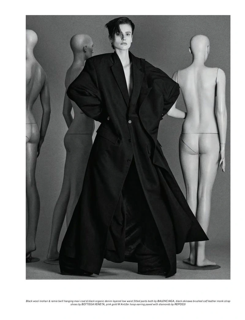 Model wearing an oversized black coat standing proudly among several mannequins in a studio setting for Pop Magazine