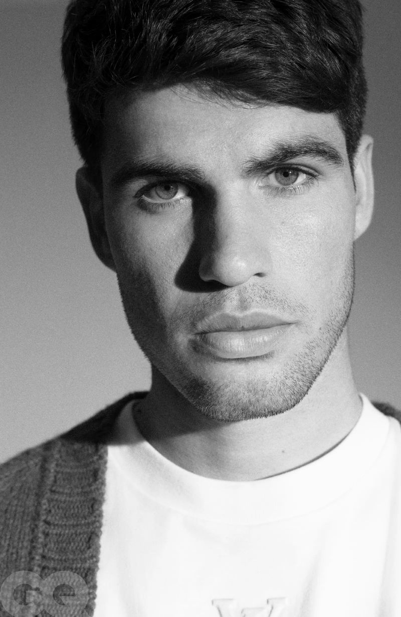 Close-up monochrome portrait of Carlos Alcaraz wearing a white t-shirt and dark cardigan under directional lighting for GQ España.