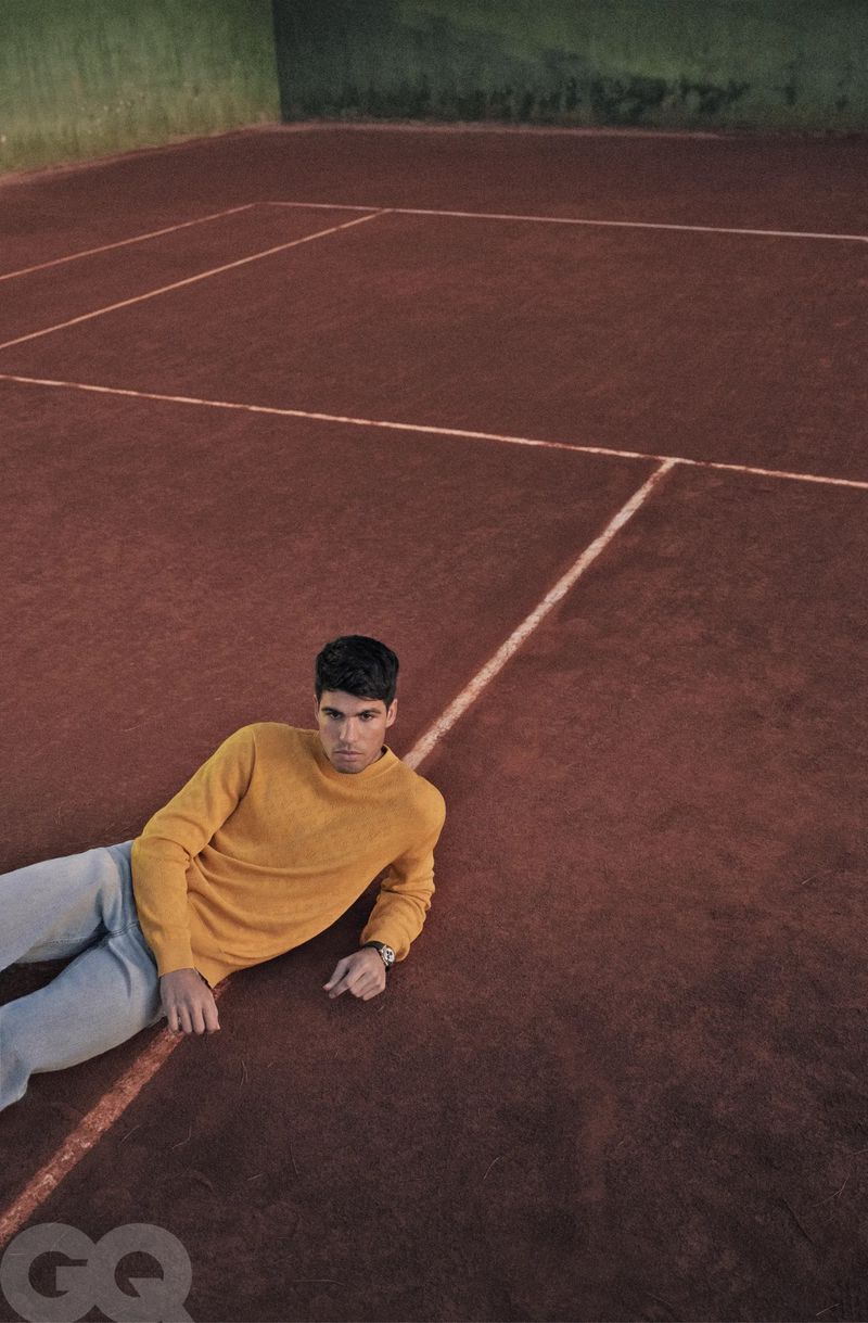 Young man in yellow sweater poses on red-clay tennis court with white lines, creating a casual fashion editorial look.