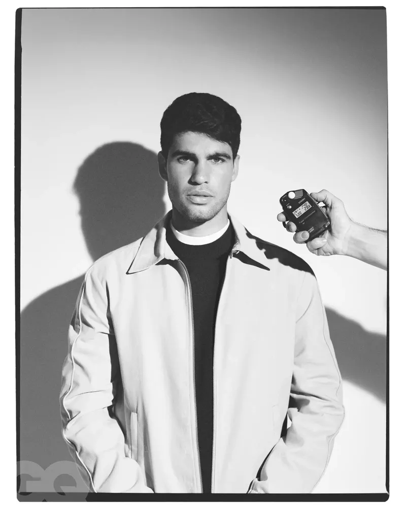 Black and white portrait of an adult man in a jacket being measured by a handheld light meter against a white background.