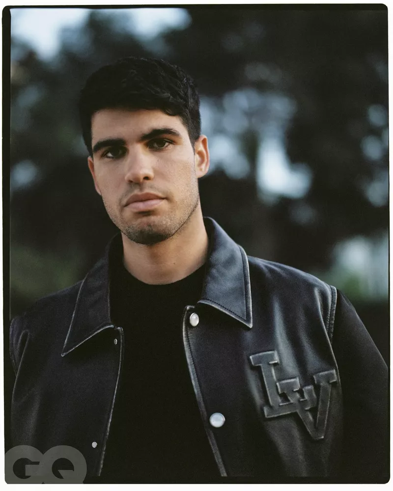 Portrait of a dark-haired adult male wearing a black jacket with the initials LV, set against a blurred outdoor background of trees.