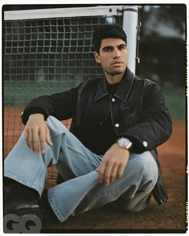Portrait of a male model sitting on a tennis court, wearing a dark jacket, light-blue jeans, and black shoes, with a tennis net in the background.
