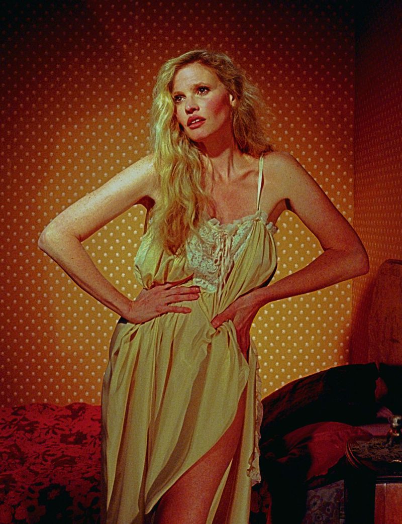 Woman with long blond hair poses in a yellow nightgown in front of a yellow and red polka dot wallpaper in an indoor setting.
