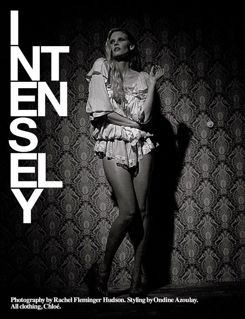 Black and white fashion shot of a slim woman in lingerie-style outfit posing against a patterned wallpaper, emphasizing intensity and style.
