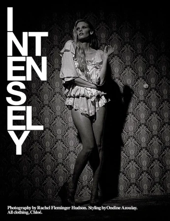 Black and white fashion shot of a slim woman in lingerie-style outfit posing against a patterned wallpaper, emphasizing intensity and style.