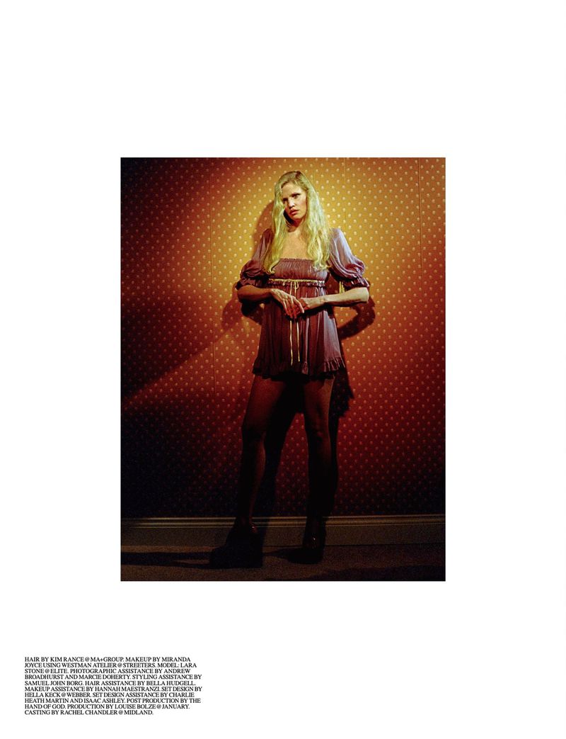 A fair-skinned woman with long blonde hair poses indoors wearing a short lilac dress and black tights, standing against a patterned wall.
