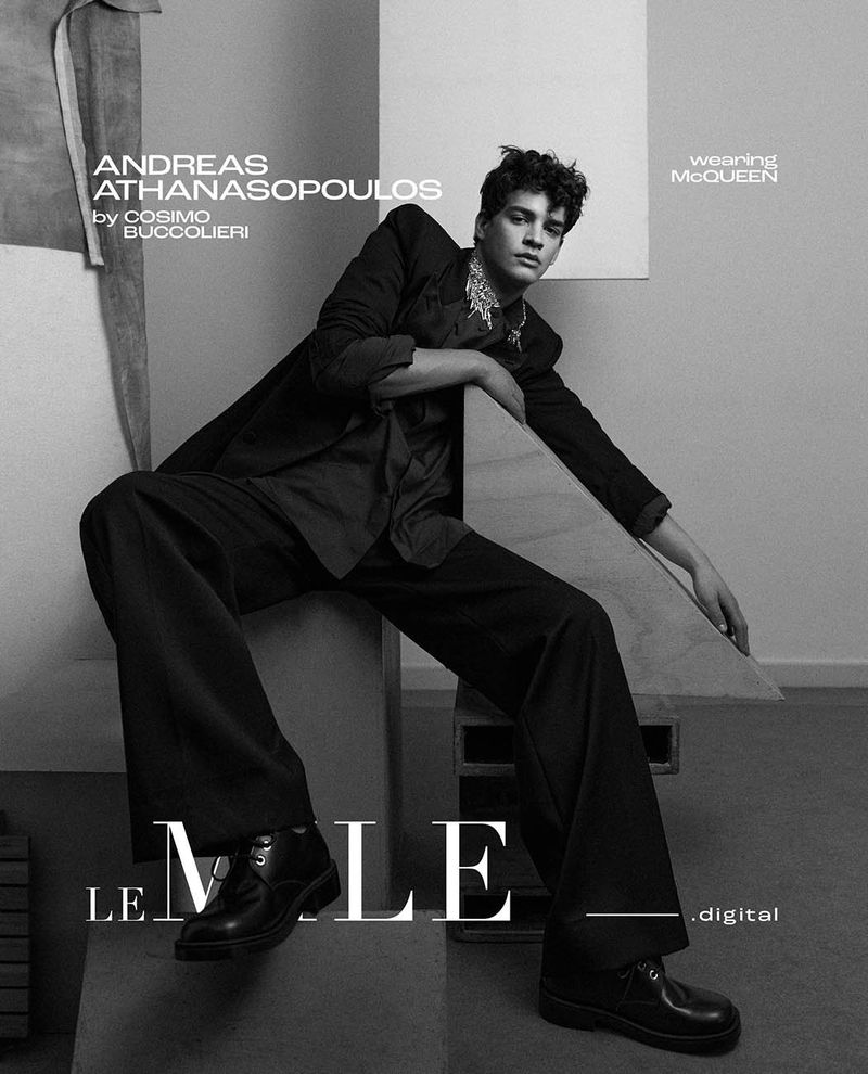 Black and white studio shot of a young adult male fashion model sitting on wooden blocks wearing a dark suit and dress shoes.