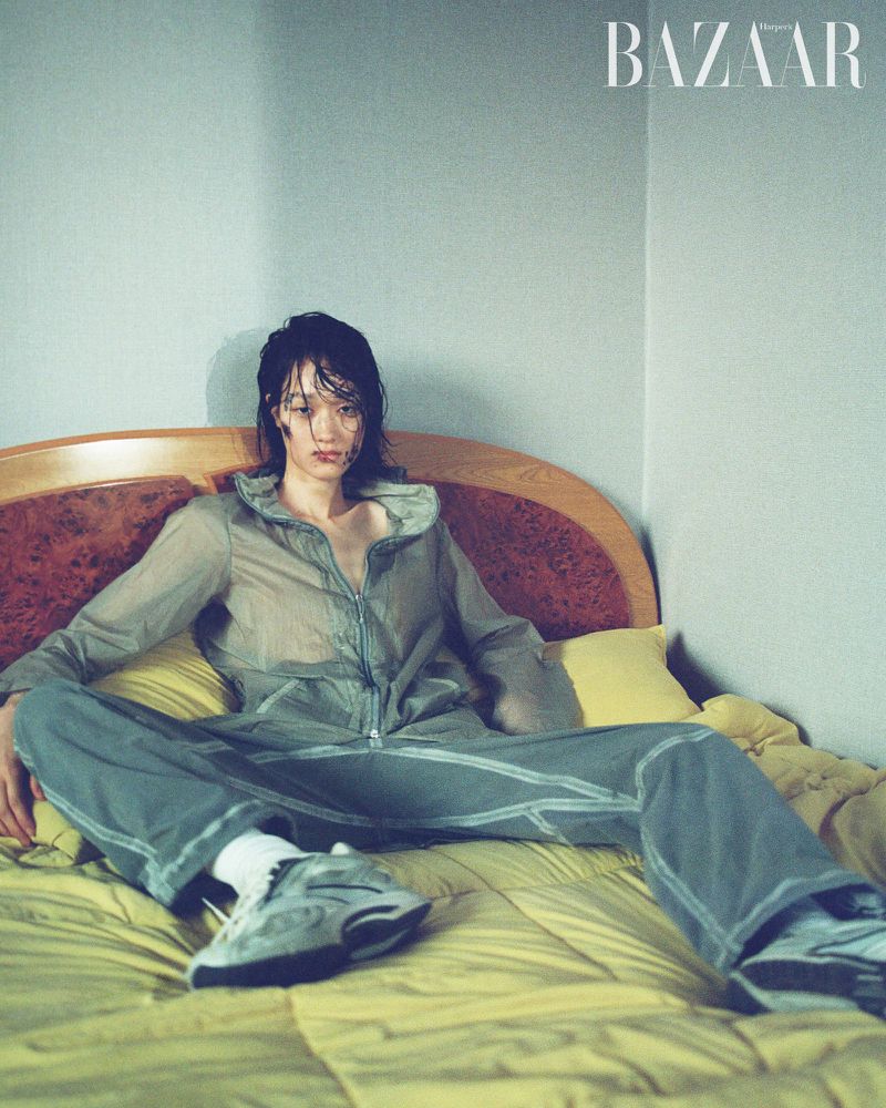 A person in a track suit sits cross-legged on a bed with a yellow quilt. The person is wearing sneakers and white socks. A magazine logo is visible.
