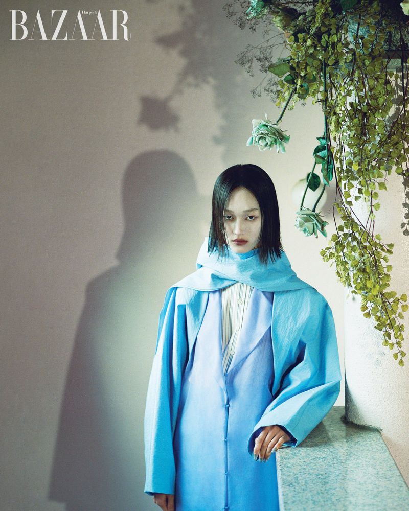 Portrait of Asian woman in a blue coat with a blue scarf, standing next to a ledge with flowers and Bazaar logo in upper left corner.