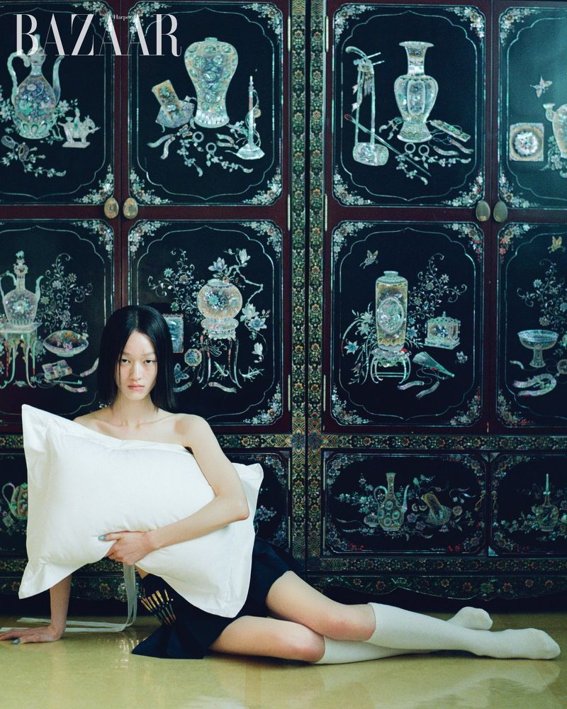 A female model poses indoors holding a white pillow, wearing a black skirt and white socks, against an ornate black and red screen.