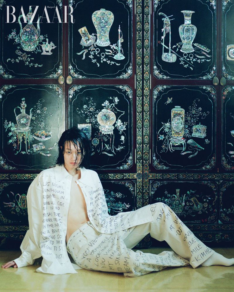 Adult male fashion model poses in patterned white shirt and pants in front of patterned dark brown folding screen indoors.
