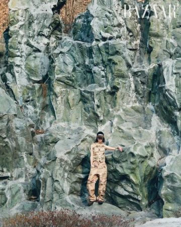 A woman in a camo outfit poses on a large rock formation with a magazine cover visible in the upper right corner on a sunny day.