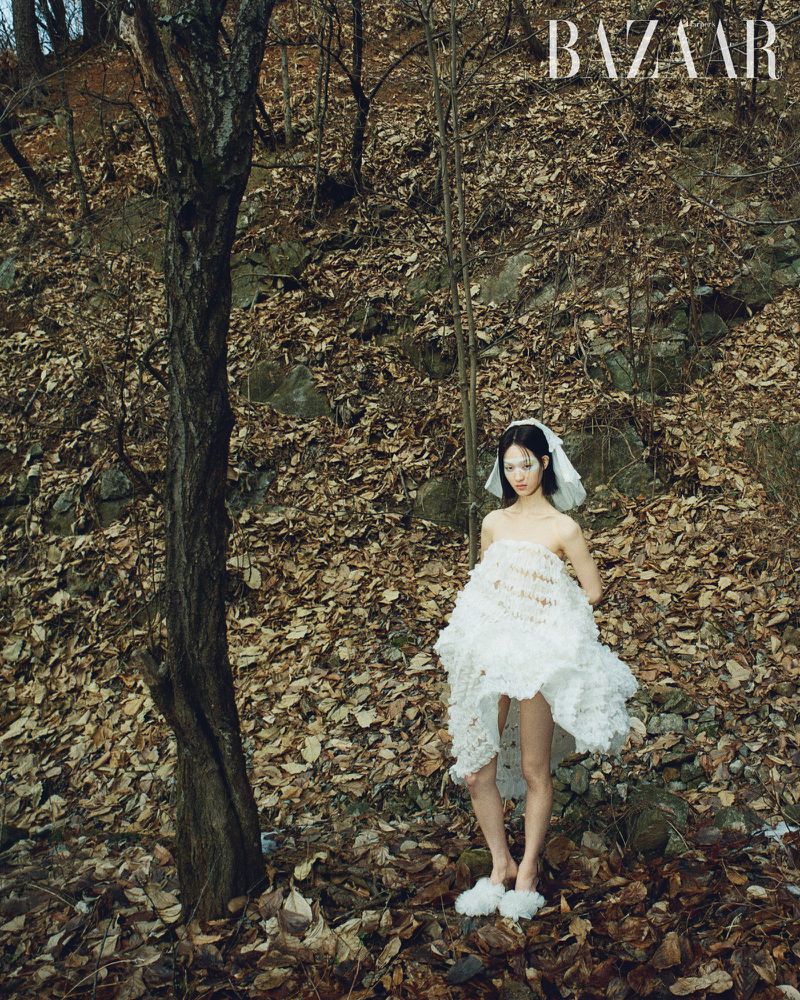Woman in nature wearing a frilly white dress stands surrounded by fallen leaves and a small tree, posing for a fashion editorial.