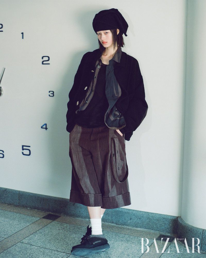 A slender young woman in oversized brown shorts, a black jacket, and a black hat stands indoors next to a wall with a clock painted on it.