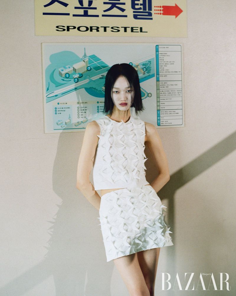 Woman posing in white geometric-patterned two-piece outfit with short hair, standing indoors in front of a sign and turquoise wall art.