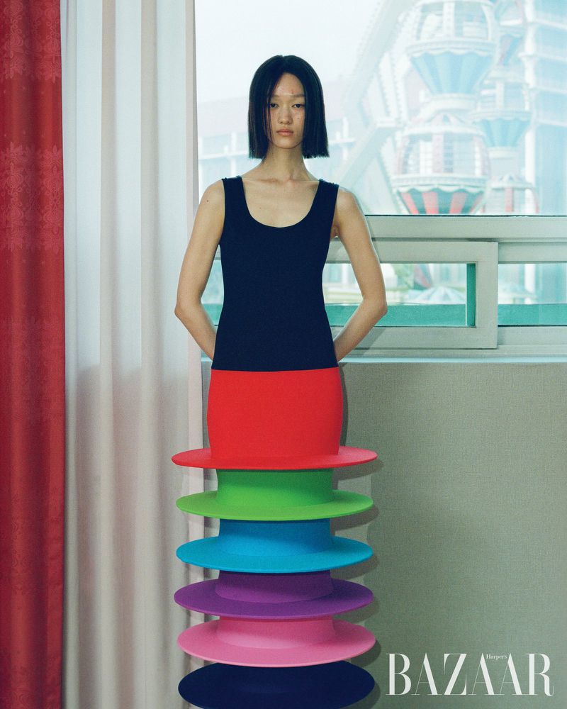 A woman with short black hair stands in front of a window, wearing a black top and a skirt made of stacked, colorful hats.