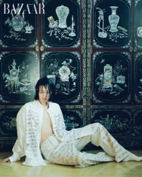 Model seated on the floor wearing printed white shirt and trousers against an ornate dark wooden cabinet background.