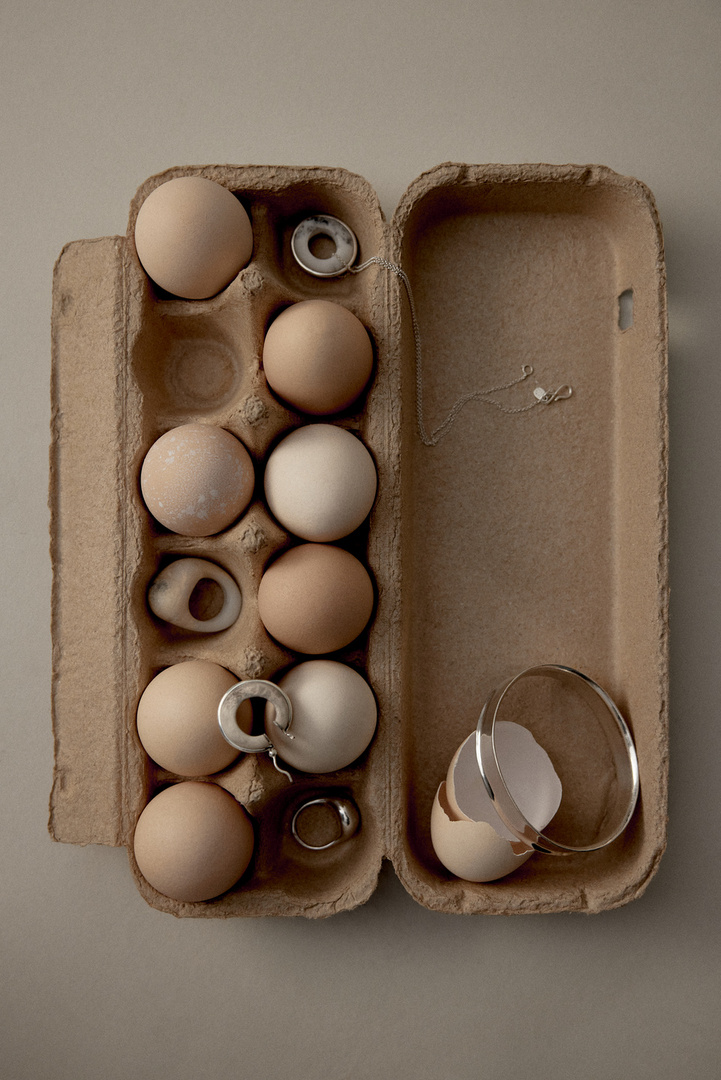 Overhead shot of a brown egg carton filled with eggs and jewelry including rings and a necklace on a neutral surface.