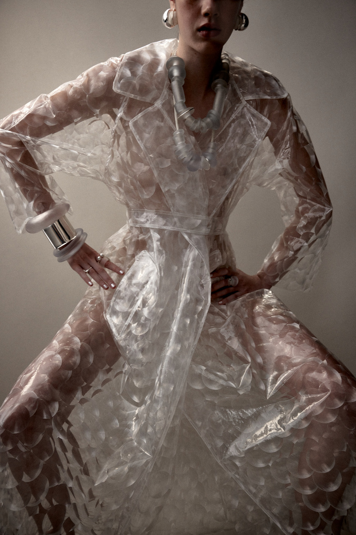 A woman in a transparent trench coat poses confidently in a studio with neutral lighting, showcasing a fashionable and artistic look.
