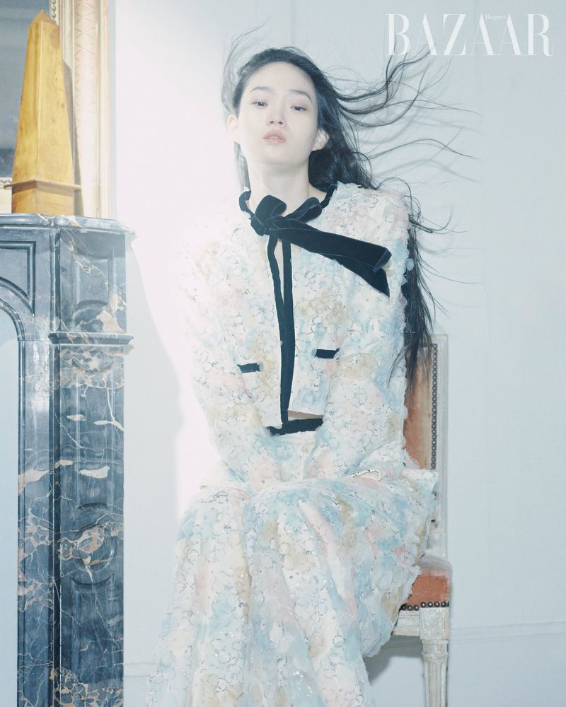 A young woman poses in a light-colored, floral-patterned dress with a black bow, seated in front of a grey marble fireplace with a mirror.