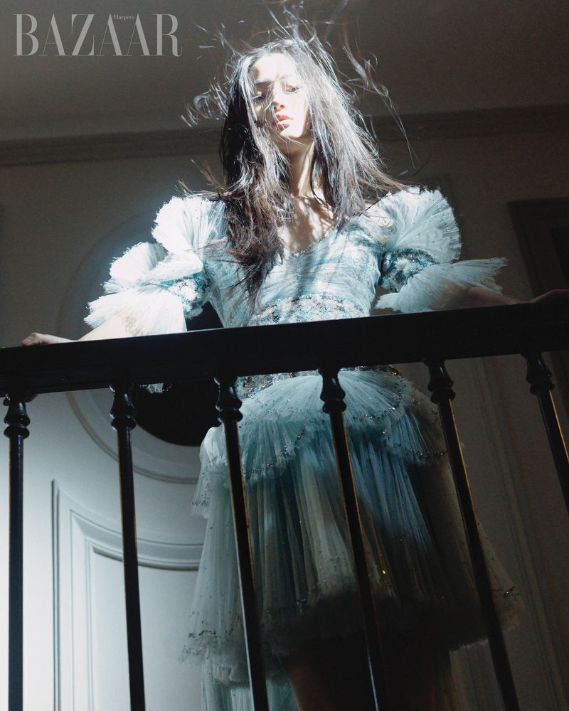 A woman in a light blue tulle dress stands on a staircase with an ornate railing, shot from a low angle with dramatic lighting.