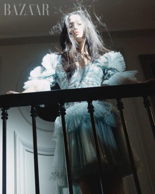 A woman in a light blue tulle dress stands on a staircase with an ornate railing, shot from a low angle with dramatic lighting.