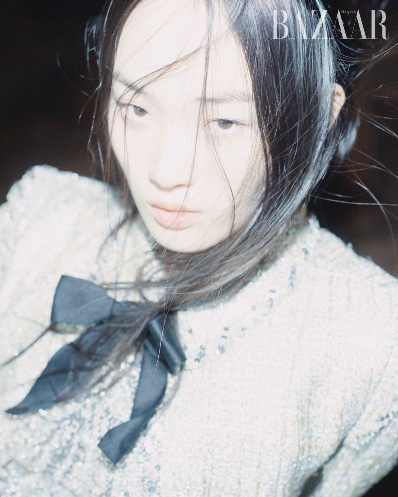 Close-up fashion shot of a young Asian woman with dark hair, wearing a sparkly white jacket with a black bow, against a dark background.