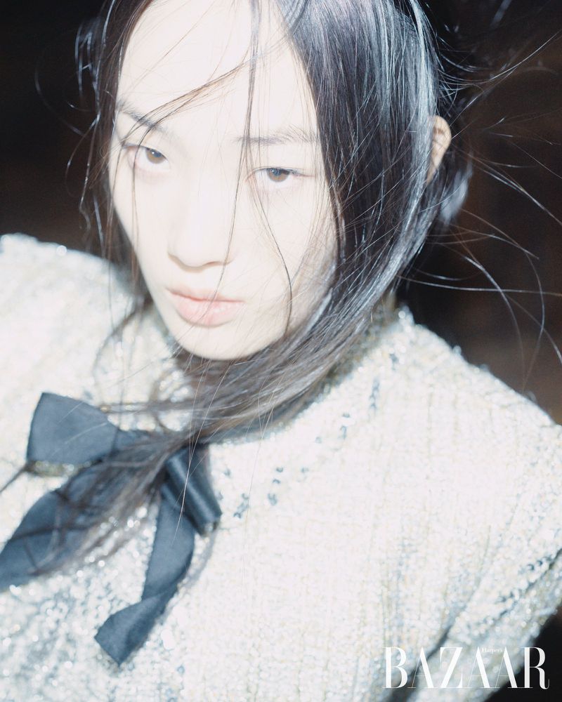 Close-up portrait of an Asian woman with fair skin and dark hair wearing a sequined jacket with a large black ribbon bow.