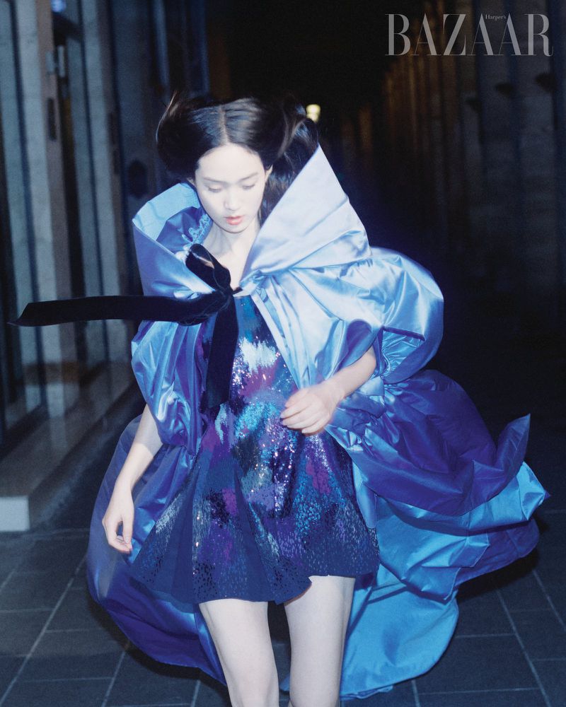 A young woman in a high-fashion photo, wearing a sparkly dress and blue cape with a large bow, against a dark urban backdrop.