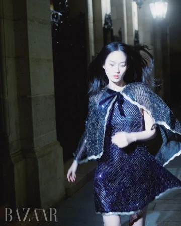 Fashion shot of a woman in a short, dark, shimmering dress with a cape, posing at night on a city street with an architectural backdrop.