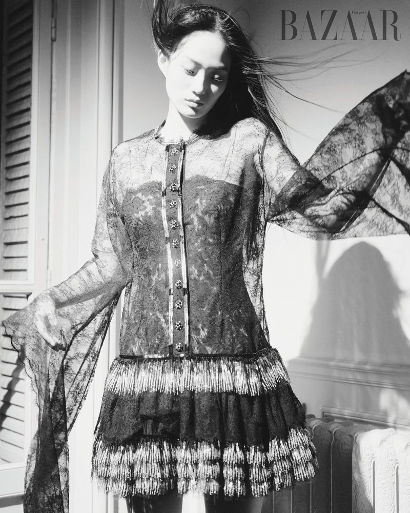 Monochrome fashion shot: Asian woman poses indoors wearing a lace dress with a tiered skirt. 'BAZAAR' logo visible in upper right corner.