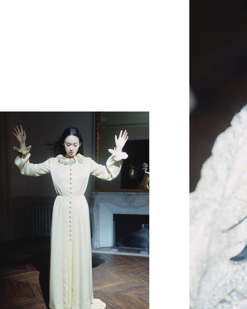Woman in a long cream gown with frilled sleeves poses with arms raised in a room with parquet floor, fireplace, and mirror.