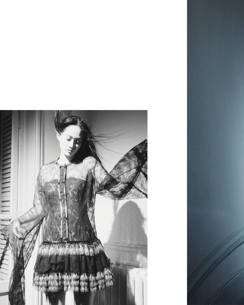 Monochrome fashion shot of a woman in an ornate black lace dress with a layered skirt, posing in a room with shuttered windows.