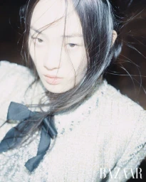 Close-up portrait of a model with dark hair blowing across her face, wearing a tweed jacket with a black bow