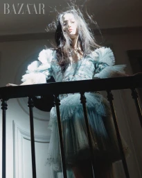 Model wearing a ruffled light blue dress leaning over a dark staircase railing under dramatic top lighting