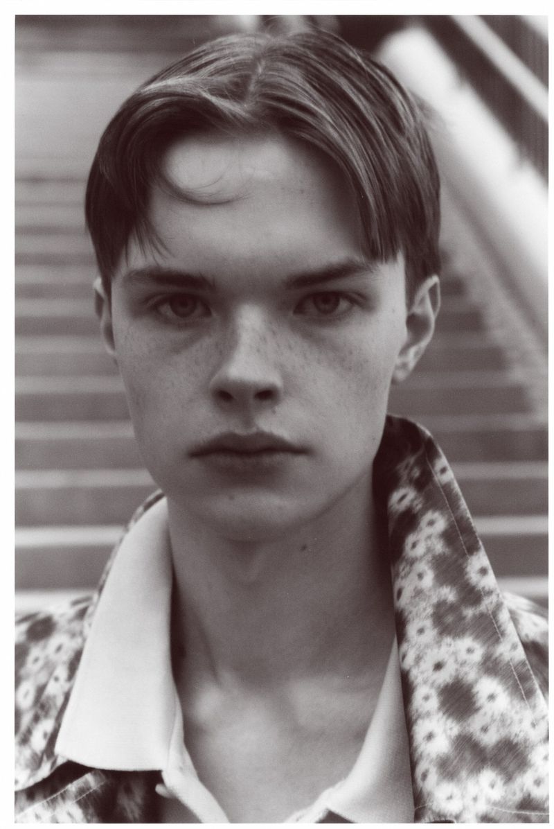 Black and white portrait of a young adult man with dark hair and light skin wearing a floral print jacket against a background of steps.