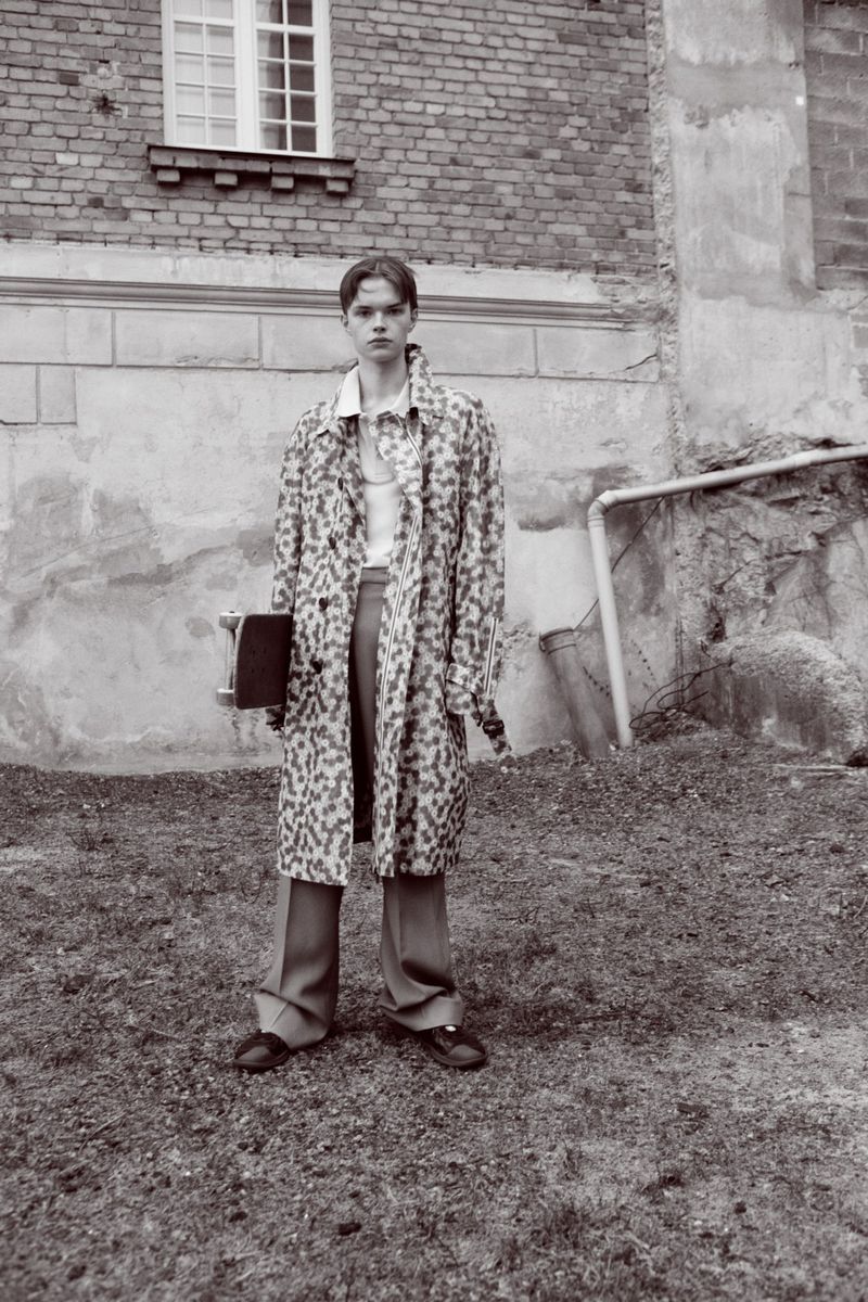 A young adult stands outdoors in a patterned coat, trousers, and shoes, holding a skateboard in front of an old brick building.