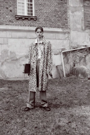 A young adult stands outdoors in a patterned coat, trousers, and shoes, holding a skateboard in front of an old brick building.