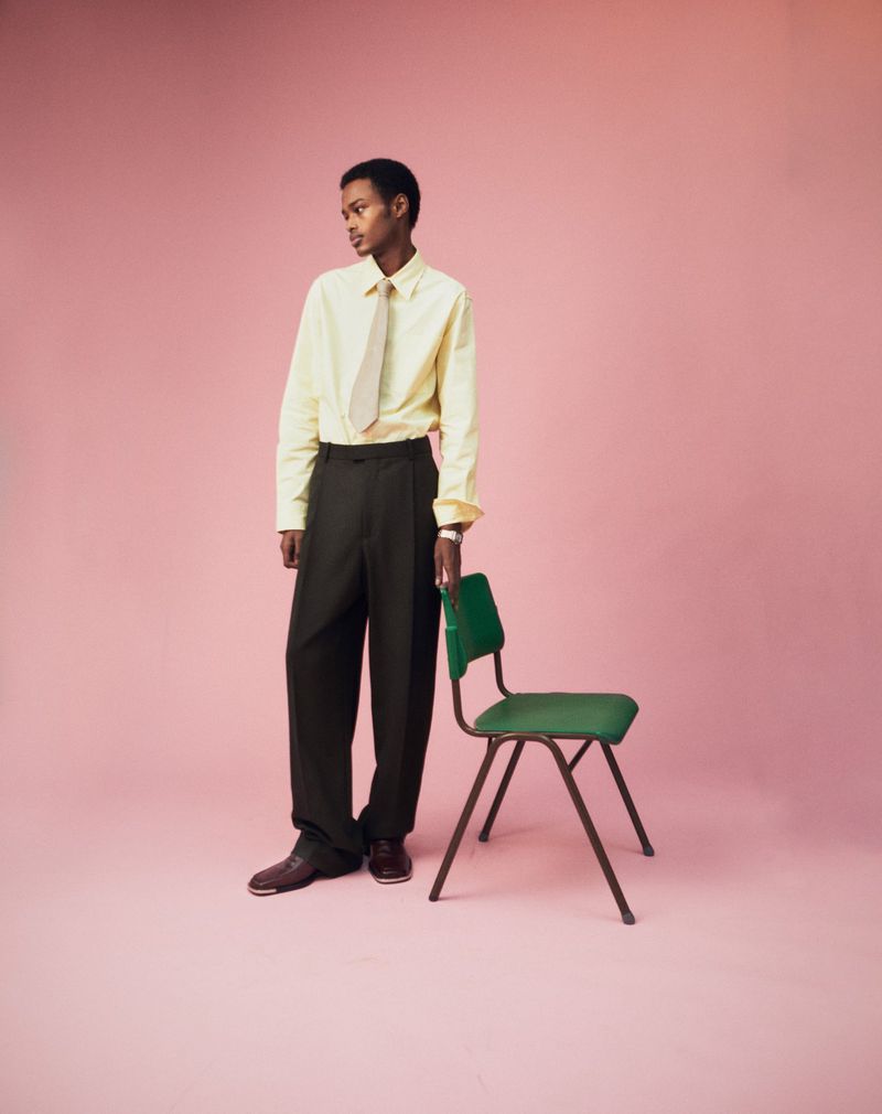 A young Black man in a dress shirt and tie stands near a green chair against a solid pink backdrop in a fashion editorial.