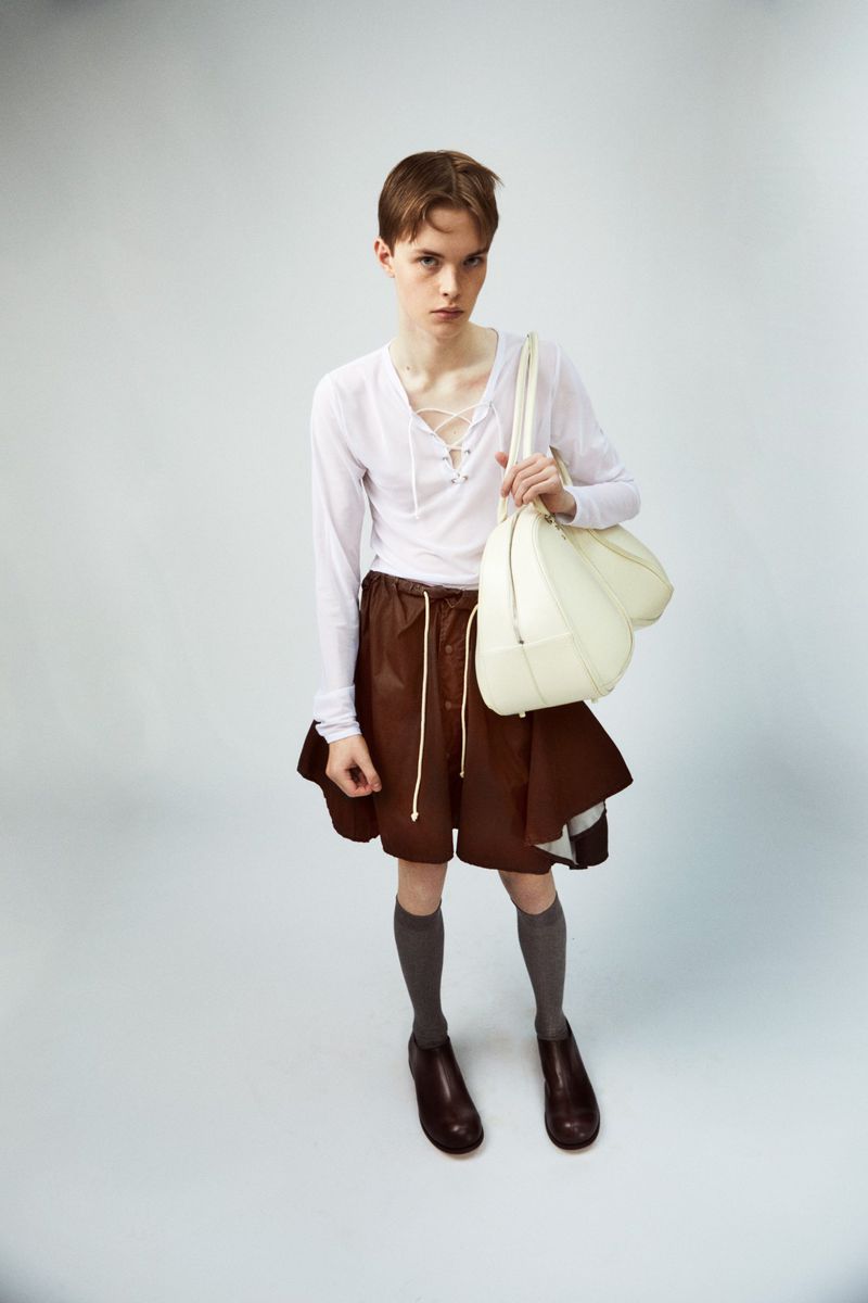 A person poses in a white long-sleeved shirt and brown shorts, carrying a white bag against a pale grey background in a studio setting.